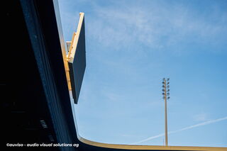 Seitliche Aufnahme des Scoreboards: Sichtbar ist das schlanke Seitenprofil der LED-Wand, ergänzt durch das Arenadach und einen Flutlichtmast im Hintergrund.