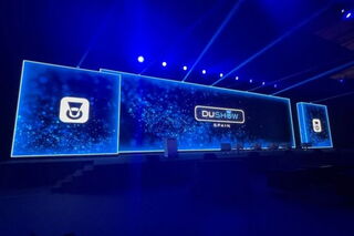 Eine Unilumin-LED-Wand auf einer Bühne installiert. Die dreiteilige Wand, deren mittlerer Teil den Hauptfokus des Bühnenbilds bildet, steht in einem blaubelichteten Konzertraum. Auf den LED-Displays sind Logos der Veranstaltung zu sehen.