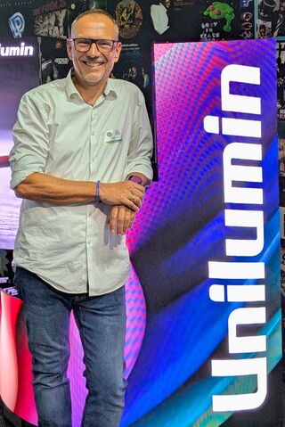 Andreas Voss Ein Bild eines Mitarbeiters der Unilumin Germany GmbH. Der Mann steht in Jeans und Hemd bekleidet vor einer LED-Wand die das Logo der Unternehmung zeigt.