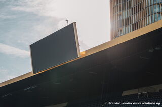 Eine Tribüne, aus nächster Nähe aufgenommen. Im Fokus steht das neu installierte LED-Scoreboard; im Hintergrund sind der blaue Himmel und ein modernes Gebäude sichtbar.