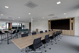 Ein modernes Bürogebäude verfügt über einen großen Konferenztisch, einen Stehtisch mit Hochstühlen, einer Couch, einem mobilen Display auf Rollen sowie einer Holzwand mit eingebauter LED-Display-Technik.