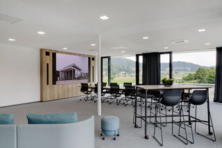 Ein modernes helles Bürogebäude von innen ausgestattet mit schwarzen Stühlen, einem Stehtisch und einem großen Konferenztisch beide aus Holz. Direkt neben der weitläufigen Glasfront wurde in einem Holzschrank ein LED-Display verbaut.