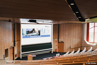 Ein Hörsaal einer Universität mit Holz verkleidet. Neben den Stufensitzen sind zwei Tafelboards erkennbar. Darüber hängt eine große LED-Wand die eine Startseite einer Präsentation zeigt.