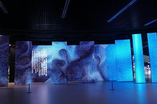 In einem dunklen Raum wird eine Kunstinstallation aus mehreren quadratischen Säulen in unterschiedlichen Größen ausgestellt. Die Säulen bestehen aus LED-Displays und zeigen Bilder in verschiedenen Blautönen.