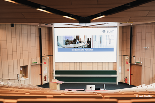 Ein Hörsaal einer Universität mit Holz verkleidet. Neben den Stufensitzen sind zwei Tafelboards erkennbar. Darüber hängt eine große LED-Wand die eine Startseite einer Präsentation zeigt.