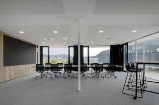 Ein modernes Bürogebäude mit zweiseitiger Glasfront und tollem Ausblick über die Stadt. Zentral im Raum befindet sich ein großer Konferenztisch mit vielen schwarzen Stühlen. Am linken Ende des Tisches befindet sich ein heller Holzschrank mit LED-Display.