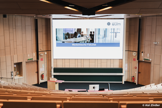 Ein Hörsaal einer Universität mit Holz verkleidet. Neben den Stufensitzen sind zwei Tafelboards erkennbar. Darüber hängt eine große LED-Wand die eine Startseite einer Präsentation zeigt.