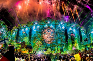 Eine Aufnahme der Hauptbühne des Tomorrowland-Festivals in Brasilien. Im Vordergrund steht eine große Menge Festivalbesucher, die Bühne verfügt über große LED-Wände mit grünen, fantasievollen Bildern. Im Hintergrund steigen Feuerwerkskörper in den Himmel.